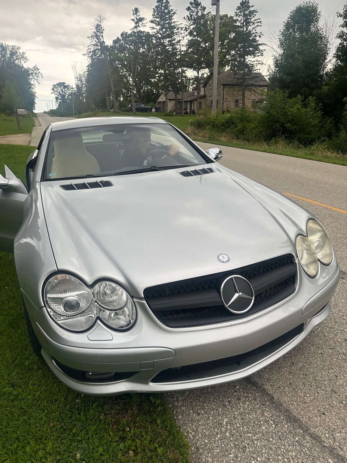 Silver Mercedes-Benz convertible parked on grass beside a rural road with trees and an old building in the background
